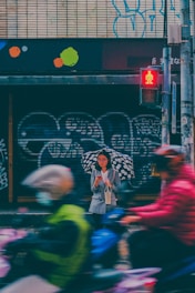 A person with glasses stands under a patterned umbrella on a rainy street. They are holding a phone and a small bag, while wearing a gray coat. Blurred motion of cyclists with colorful jackets and helmets passes by, and a red pedestrian crossing signal is visible. The background features graffiti and colorful shapes on a tiled wall.