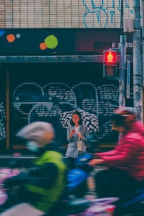 A person with glasses stands under a patterned umbrella on a rainy street. They are holding a phone and a small bag, while wearing a gray coat. Blurred motion of cyclists with colorful jackets and helmets passes by, and a red pedestrian crossing signal is visible. The background features graffiti and colorful shapes on a tiled wall.