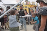 A group of people, including children and adults, are participating in an outdoor activity involving foam swords and protective gear. They are surrounded by metal scaffolding and various structures, with some individuals appearing to be cheering or watching intently.