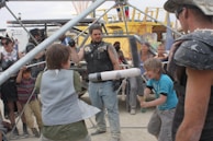 A group of people, including children and adults, are participating in an outdoor activity involving foam swords and protective gear. They are surrounded by metal scaffolding and various structures, with some individuals appearing to be cheering or watching intently.