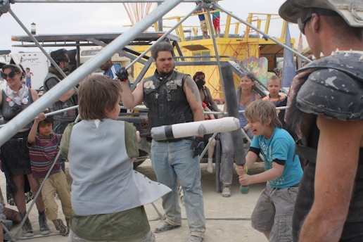 A group of people, including children and adults, are participating in an outdoor activity involving foam swords and protective gear. They are surrounded by metal scaffolding and various structures, with some individuals appearing to be cheering or watching intently.