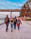 Four people with colorful hair and backpacks walk along a riverside promenade. The scene features a bridge spanning over the walkway, with a few vehicles on it. Leafy trees line the path on one side, and the river is visible on the other. The setting suggests a leisurely outing in an urban park area.
