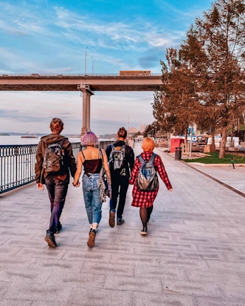 Four people with colorful hair and backpacks walk along a riverside promenade. The scene features a bridge spanning over the walkway, with a few vehicles on it. Leafy trees line the path on one side, and the river is visible on the other. The setting suggests a leisurely outing in an urban park area.