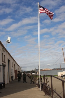 Several people in military uniforms stand near a large flagpole displaying the American flag. The setting is an industrial area with warehouses and a parking lot. The sky is partly cloudy, and there is a surveillance camera mounted on the building.