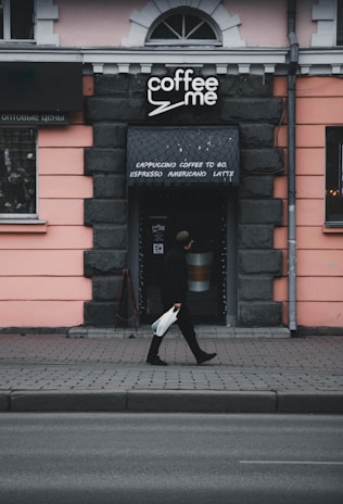 A person walks past a small coffee shop with a sign that reads 'coffee me'. The shop is set in a building with a light pink facade and dark stone-like trim. The entrance has a small awning listing types of coffee, such as cappuccino and espresso. The scene is slightly overcast, contributing to a somewhat muted color palette.