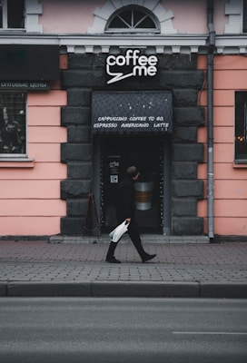 A person walks past a small coffee shop with a sign that reads 'coffee me'. The shop is set in a building with a light pink facade and dark stone-like trim. The entrance has a small awning listing types of coffee, such as cappuccino and espresso. The scene is slightly overcast, contributing to a somewhat muted color palette.