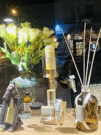 A warmly lit shop window display features a variety of home decor items. On the left, there's an ornate vase with pale yellow flowers. In front are jars and bottles labeled with the brand 'Zielinski & Rozen', a candle on a gold stand, and a horse-head figurine. To the right, a large amber bottle with white reeds sticks is displayed.