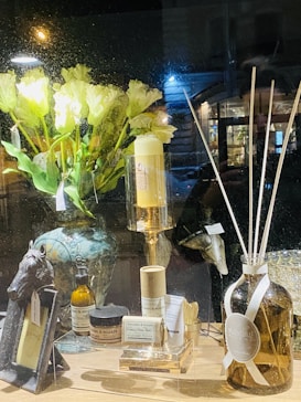 A warmly lit shop window display features a variety of home decor items. On the left, there's an ornate vase with pale yellow flowers. In front are jars and bottles labeled with the brand 'Zielinski & Rozen', a candle on a gold stand, and a horse-head figurine. To the right, a large amber bottle with white reeds sticks is displayed.