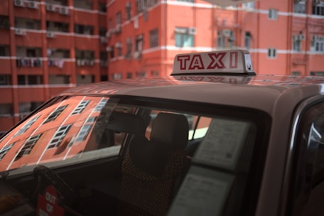 A taxi with a prominent roof sign is parked in front of a red brick apartment building. The windows of the building reflect sunlight, and laundry can be seen hanging from balconies.
