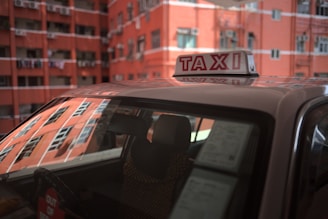A taxi with a prominent roof sign is parked in front of a red brick apartment building. The windows of the building reflect sunlight, and laundry can be seen hanging from balconies.