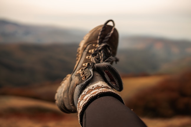 A single hiking boot rests atop a blurred mountain landscape backdrop. The boot is rugged and well-worn, paired with a thick, patterned sock. The background shows earthy tones of the mountains, suggesting a serene and possibly autumn setting.