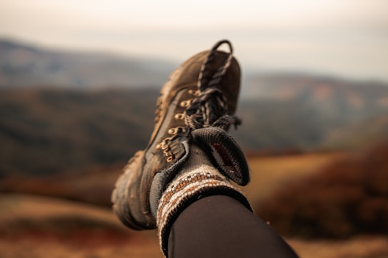 A single hiking boot rests atop a blurred mountain landscape backdrop. The boot is rugged and well-worn, paired with a thick, patterned sock. The background shows earthy tones of the mountains, suggesting a serene and possibly autumn setting.