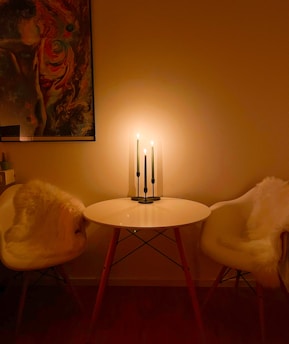 A cozy, softly-lit room with a small, round white table placed between two white fur-draped chairs. Three tall candles on a black stand provide a warm glow, adding to the intimate ambiance. A colorful abstract painting is partially visible on the wall.