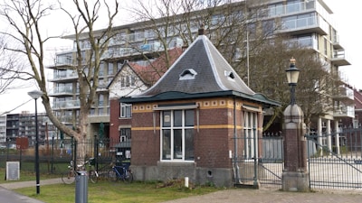 A small brick building with a slate roof and decorative brickwork stands in front of a modern apartment block. The building is surrounded by a metal fence with a narrow road beside it. Leafless trees arch over the scene, casting intricate shadows. Two bicycles are parked near the fence, and an old-fashioned street lamp is nearby.