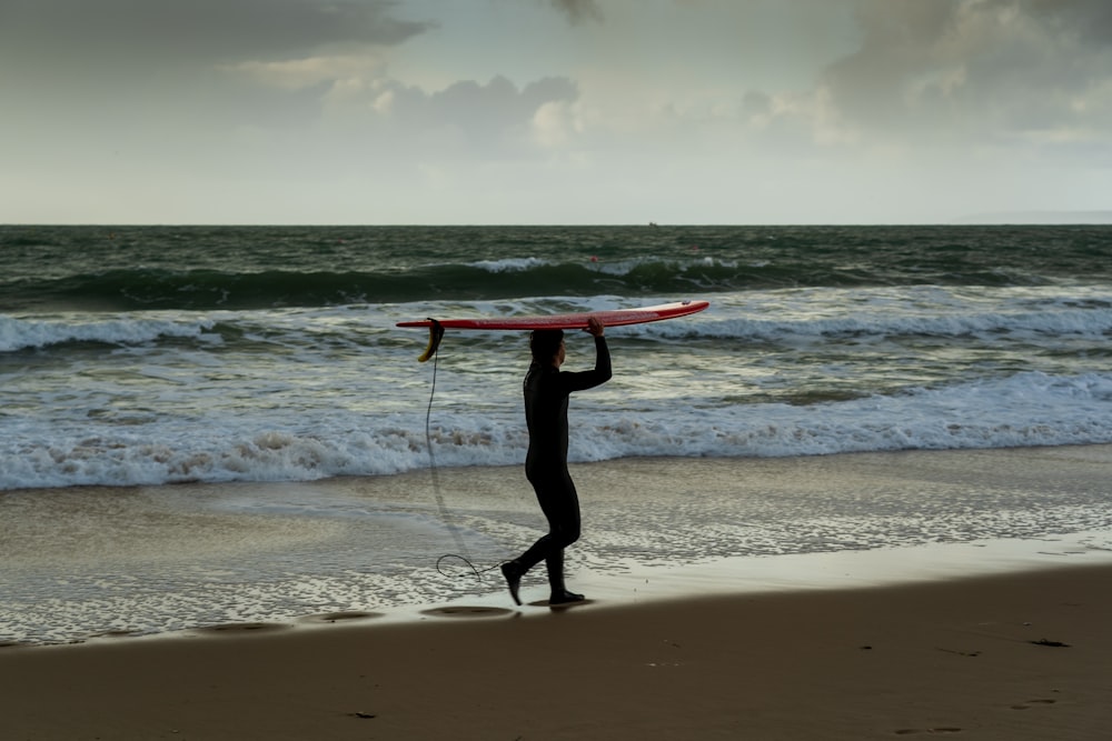 man in black wet suit holding red surfboard standing on beach during daytime