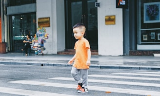 A young child wearing an orange shirt and gray pants is walking across a zebra crossing on a city street. In the background, there is a seated woman with a cart filled with colorful items, likely a street vendor, and some storefronts.