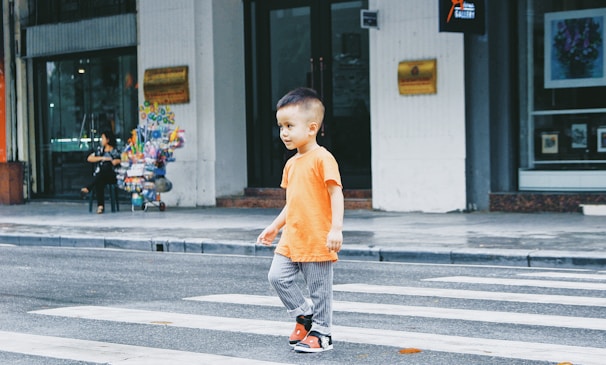 A young child wearing an orange shirt and gray pants is walking across a zebra crossing on a city street. In the background, there is a seated woman with a cart filled with colorful items, likely a street vendor, and some storefronts.
