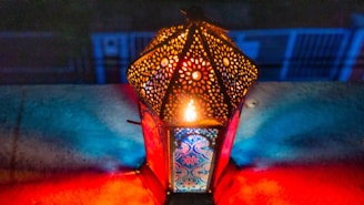 An intricately designed, illuminated lantern emits colorful light patterns across a surface. The lantern has a lattice design, allowing the light to create a vivid display of red and blue hues in its surroundings.