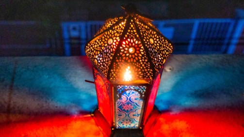 An intricately designed, illuminated lantern emits colorful light patterns across a surface. The lantern has a lattice design, allowing the light to create a vivid display of red and blue hues in its surroundings.