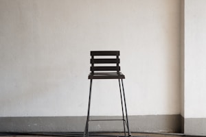 A simple wooden chair with a backrest made of horizontal slats stands against a plain white wall. The chair has long metal legs and is positioned on a floor with a neutral tone.