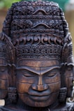 Intricately carved wooden statue featuring a detailed and ornate head with a serene facial expression. The craftsmanship highlights elaborate patterns and textures, reflecting cultural artistry.