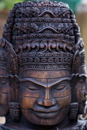 Intricately carved wooden statue featuring a detailed and ornate head with a serene facial expression. The craftsmanship highlights elaborate patterns and textures, reflecting cultural artistry.