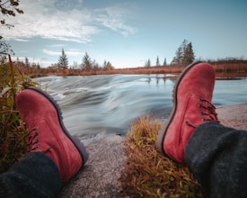 A pair of red boots is prominently displayed in the foreground, resting on a rock near a flowing river. The water appears in motion, creating a smooth visual effect. Grass and small plants grow beside the rock. The background features tall evergreen trees under a bright blue sky with scattered clouds.