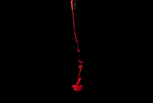 A stream of bright red liquid is flowing down through a dark black background, creating an abstract, dramatic effect.
