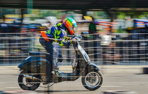 A person wearing a colorful helmet and protective gear races on a scooter. The background is blurred, indicating speed and motion. The setting appears to be an outdoor track with onlookers behind barriers.