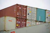 Multiple freight containers are stacked in an organized manner, featuring colors like red, blue, and beige. They have various shipping labels and logos, with caution signs present on some containers.