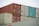 Multiple freight containers are stacked in an organized manner, featuring colors like red, blue, and beige. They have various shipping labels and logos, with caution signs present on some containers.
