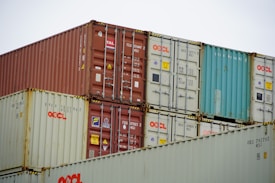 Multiple freight containers are stacked in an organized manner, featuring colors like red, blue, and beige. They have various shipping labels and logos, with caution signs present on some containers.