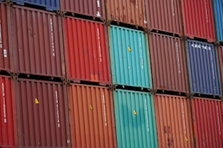 A stacked arrangement of intermodal shipping containers, featuring a variety of colors including red, blue, and teal. Each container has distinct numbering and labeling, with some visible brand names and caution symbols.