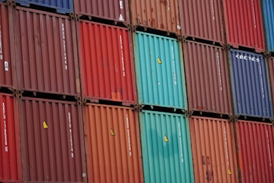 A stacked arrangement of intermodal shipping containers, featuring a variety of colors including red, blue, and teal. Each container has distinct numbering and labeling, with some visible brand names and caution symbols.