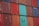 A stacked arrangement of intermodal shipping containers, featuring a variety of colors including red, blue, and teal. Each container has distinct numbering and labeling, with some visible brand names and caution symbols.