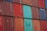 A stacked arrangement of intermodal shipping containers, featuring a variety of colors including red, blue, and teal. Each container has distinct numbering and labeling, with some visible brand names and caution symbols.