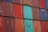 A stacked arrangement of intermodal shipping containers, featuring a variety of colors including red, blue, and teal. Each container has distinct numbering and labeling, with some visible brand names and caution symbols.