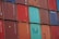 A stacked arrangement of intermodal shipping containers, featuring a variety of colors including red, blue, and teal. Each container has distinct numbering and labeling, with some visible brand names and caution symbols.