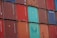 A stacked arrangement of intermodal shipping containers, featuring a variety of colors including red, blue, and teal. Each container has distinct numbering and labeling, with some visible brand names and caution symbols.