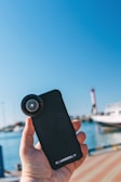 A hand is holding a smartphone with a lens attachment. The background features a clear blue sky and a blurred view of a marina with a boat and lighthouse.