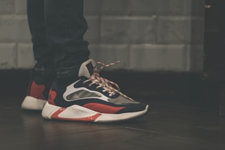A close-up view of a person wearing stylish sneakers with a striking design, combining dark blue, red, and white colors. The shoes have a textured, thick sole and patterned laces. The person is wearing dark jeans, and the background features a tiled wall with a muted color scheme.