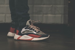 A close-up view of a person wearing stylish sneakers with a striking design, combining dark blue, red, and white colors. The shoes have a textured, thick sole and patterned laces. The person is wearing dark jeans, and the background features a tiled wall with a muted color scheme.