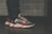 A close-up view of a person wearing stylish sneakers with a striking design, combining dark blue, red, and white colors. The shoes have a textured, thick sole and patterned laces. The person is wearing dark jeans, and the background features a tiled wall with a muted color scheme.