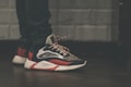 A close-up view of a person wearing stylish sneakers with a striking design, combining dark blue, red, and white colors. The shoes have a textured, thick sole and patterned laces. The person is wearing dark jeans, and the background features a tiled wall with a muted color scheme.