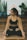 A woman is sitting in a yoga pose on a mat indoors. She is wearing a black sports bra and black leggings. The background includes various potted plants and a logo or sign that says 'Yoga'. The setting appears calm and focused on fitness or wellness.