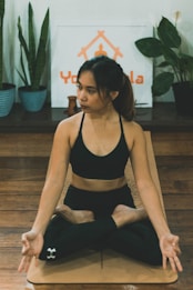 A woman is sitting in a yoga pose on a mat indoors. She is wearing a black sports bra and black leggings. The background includes various potted plants and a logo or sign that says 'Yoga'. The setting appears calm and focused on fitness or wellness.