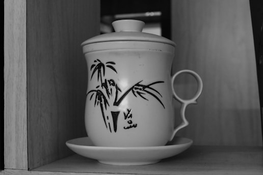 A ceramic mug with a fitted lid and a handle, adorned with a painted design of bamboo and abstract symbols, placed on a matching saucer in a wooden shelf.