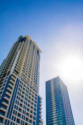 Two modern skyscrapers rise against a clear blue sky, with the sun shining brightly in the background. The buildings feature a grid-like facade with numerous windows, and one is labeled 'Lakeside Residence.'