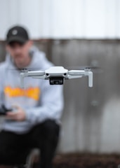 A close-up of a white drone in flight with blurred blades, centered in the foreground. In the background, a person wearing a black cap and gray hoodie appears softly out of focus, holding a controller.