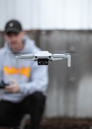 A close-up of a white drone in flight with blurred blades, centered in the foreground. In the background, a person wearing a black cap and gray hoodie appears softly out of focus, holding a controller.